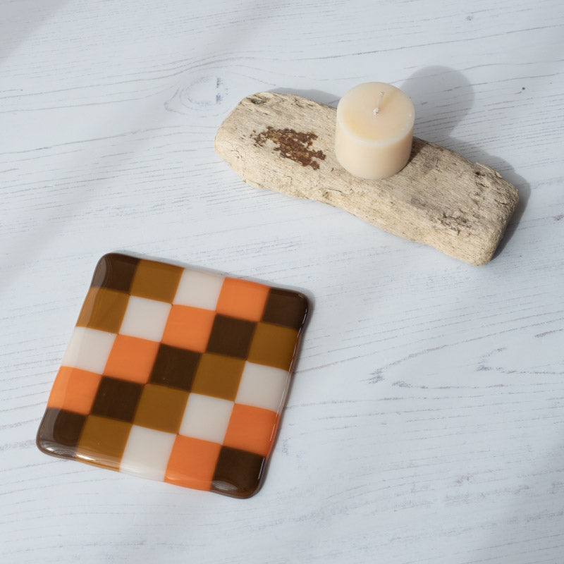 Handmade checkered glass coaster in orange, brown, and white, styled with driftwood and candle on white wood